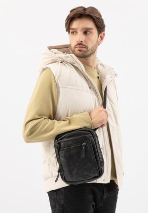 Man met kort bruin haar, gekleed in een beige vest en een olijfgroene trui, met een zwarte leren schoudertas over zijn schouder.