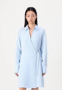 Vestito avvolgente azzurro chiaro con maniche lunghe e colletto. Tessuto strutturato, presenta una tasca sul petto e una cintura regolabile in vita.