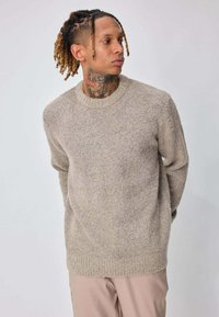 Homme avec des dreadlocks et un tatouage au cou portant un pull en maille beige et un pantalon de couleur claire, regardant vers sa droite avec les mains derrière le dos.