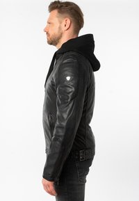 Giacca in pelle nera con cappuccio, chiusura con zip sul davanti e tasche laterali. Include dettagli su spalle e una vita con cinturino.