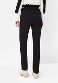 Zwarte rechte broek met een gladde textuur, een taille die net onder de navel valt en twee achterzakken. Gecombineerd met stevige lichtgekleurde schoenen.