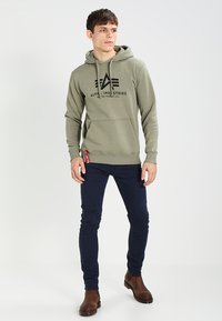 Olivegrüner Kapuzenpullover mit schwarzem Logo, Kordelzugkapuze, Kängurutasche und gerippten Bündchen. Kombiniert mit dunkelblauen Slim-Fit-Hosen und braunen Stiefeln.