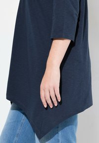 Mörkblå tunika med trekvartslång ärm, något textil med en böjd nederkant. Hand synlig som vilar på tyget.