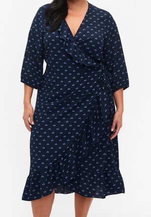 Frau trägt ein marineblaues Wickelkleid mit kleinen blauen Kirschmotiven, Rüschensaum, dreiviertel Ärmeln und seitlicher Bindung.