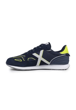 Zapatilla azul marino con materiales de ante y malla, que presenta acentos blancos y un talón amarillo brillante. Diseño con cordones y detalles perforados.