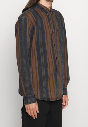 Personne portant une chemise à manches longues boutonnée avec des rayures verticales marron et bleu foncé et un pantalon noir sur un fond uni.