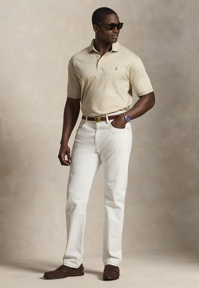 Homme portant un polo beige, un jean blanc, une ceinture et des chaussures marron, une montre bleue et des lunettes de soleil, debout avec une main dans la poche.