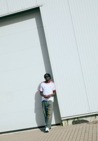 Vit idrottströja med röda detaljer och logotyp, grå denimjeans, svarta solglasögon, stående mot en ljusgrå vägg med skuggor.