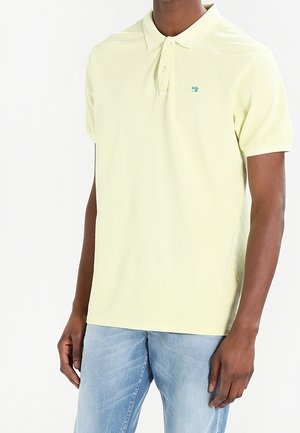 Mężczyzna ubrany w jasnożółtą koszulkę polo z krótkim rękawem i jasnoniebieskie dżinsy, stojący na białym tle.