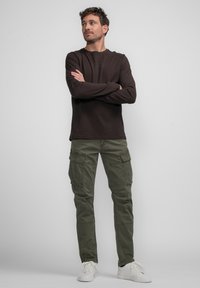 Chemise marron à manches longues avec logo en relief, associée à un pantalon cargo vert olive comportant plusieurs poches. Des baskets blanches complètent le look.