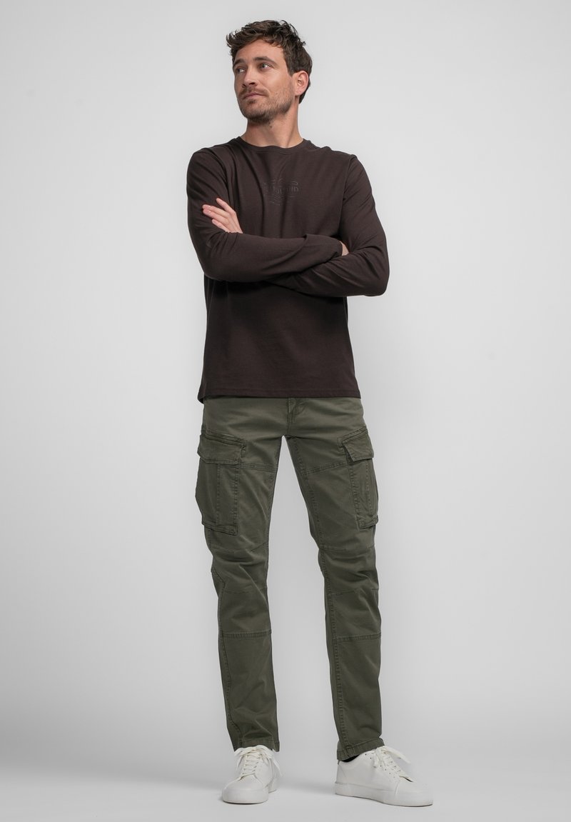Chemise marron à manches longues avec logo en relief, associée à un pantalon cargo vert olive comportant plusieurs poches. Des baskets blanches complètent le look.