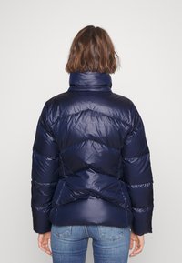 Mörkblå pufferjacka med hög krage, chevronmönster och glänsande yta. Har horisontell quiltning och elastiska ärmslut.