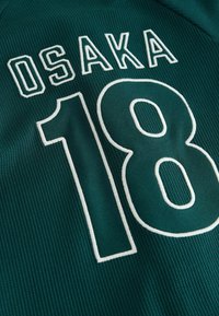 Zelený športový dres s bielym vyšívaným nápisom "OSAKA" nad číslom "18". Materiál má textúrovaný povrch.