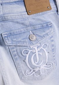 Τσέπη σε ανοιχτό μπλε τζιν με λευκό κεντητό λογότυπο TJC, ασημί κουμπί, μικρές καρφίτσες και καφέ δερμάτινη ετικέτα Juicy Couture από πάνω.