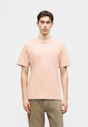 Junger Mann mit kurzen braunen Haaren, der ein hellrosa kurzärmeliges T-Shirt und hellbraune Hosen trägt, vor einem schlichten Hintergrund stehend.