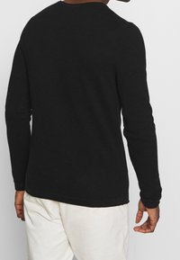 Schwarzer, strukturierter Langarmpullover mit rundem Ausschnitt, der eine figurnahe Passform und minimalistischen Stil aufweist. Weiße Hosen sind teilweise sichtbar.