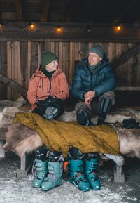 Botas de nieve en azul verdoso con detalles en rosa, al lado de un edredón amarillo en un banco de madera. Dos personas con ropa de outdoor se sientan contra un fondo de madera.