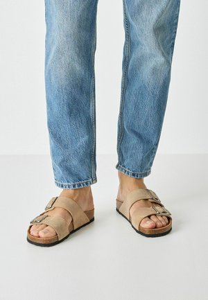 Beige ruskindsandaler med to justerbare remme og sølvspænder, kombineret med lyseblå denimjeans. Sålen er mørk og tekstureret.