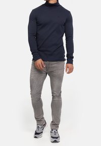 Homme portant un col roulé bleu marine, un jean gris clair et des baskets grises, bleu marine et blanches, debout devant un fond blanc uni.