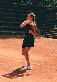 Černé sportovní šaty s červenými akcenty, bílé a fialové boty, držící tenisovou raketu na antukovém kurtu, s kamennými schody v pozadí.