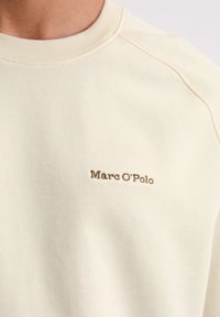 Tricou dintr-o țesătură ușoară de crem, cu guler rotund și mâneci raglan. Prezintă un logo brodat subtil "Marc O'Polo" de culoare maro pe piept.