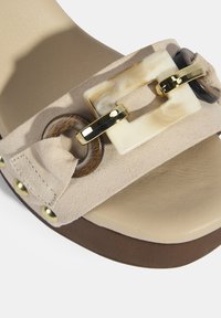 Beige Wildledersandale mit einem auffälligen rechteckigen cremfarbenen Akzent, kreisförmigen Holzringen und goldenen Beschlägen auf einer glatten, hellen Sohle.