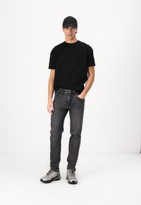 Svart T-skjorte, mørkegrå jeans, grå sneakers. Modellen står rett opp og ned, iført en svart caps. Enkel design, nøytrale farger, avslappet passform.