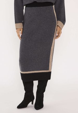 Femme portant une jupe midi en maille gris foncé avec des bordures beige et noires, associée à des bottes hautes noires à talons, sur un fond blanc.