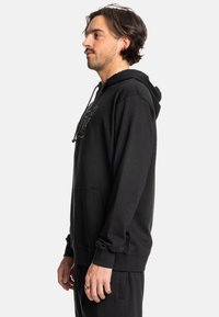 Sudadera negra de tejido suave, con un bolsillo frontal, cordones y un ajuste relajado. Incluye un logotipo gráfico en un tono más oscuro.