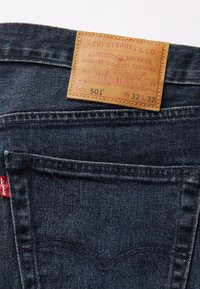 Ciemnoniebieskie dżinsy z skórzaną metką. Metka zawiera wytłoczone napisy, Lot 501 oraz wymiary W 32 L 32. Czerwona naszywka na kieszeni z detalami przeszycia.