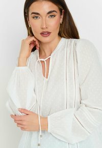 Blusa blanca con tejido texturizado, presenta un escote con lazo, mangas largas abullonadas y detalles de corte vertical a lo largo de las costuras.