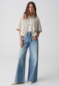 Beige oversized shirt met korte mouwen, gecombineerd met lichtblauwe wijde jeans en crèmekleurige schoenen. Denim vertoont subtiele vervaging.