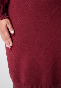 Bordeauxrode gebreide jurk met een textuur van een chevronpatroon, geribbelde boorden en een zachte afwerking, deels zichtbaar met een hand op de stof.