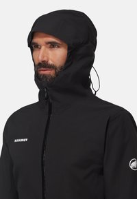 Veste noire imperméable avec un col haut et une capuche réglable, dotée d'une fermeture à glissière sur le devant et d'un accent de logo blanc sur la manche.