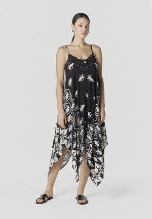 Mujer con vestido de estampado en blanco y negro con dobladillo tipo pañuelo, tirantes finos y sandalias planas negras, de pie sobre un fondo liso.