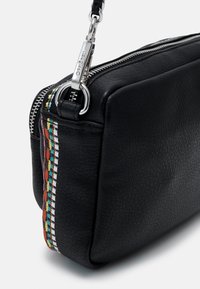 Musta nahkalaukku, jossa on teksturoitu pinta ja hopeanvärinen vetoketju. Sivussa värikkäitä kudottuja yksityiskohtia ja irrotettava hihna.