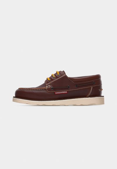 Chaussure bateau en cuir marron avec lacets jaunes, semelle blanche, œillets noirs, détail de laçage latéral, et étiquette logo rouge "DSQUARED2".