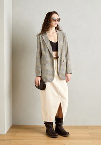 Blazer à carreaux beige et noir, crop top noir, jupe crème avec fente et bottines brunes. Les accessoires comprennent une pochette noire et des lunettes de soleil.