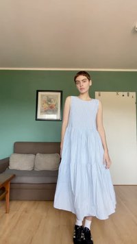 Personne aux cheveux courts portant une robe sans manches bleu clair rayée et des baskets noires, debout dans un salon aux murs verts.