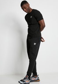 Camiseta negra de manga corta con logo blanco en el pecho, combinada con pantalones jogger negros con franjas laterales y marca en blanco. Zapatillas multicolores.