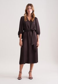 Braunes, langärmliges Kleid aus weichem Stoff, mit einem V-Ausschnitt und einem gebundenen Gürtel in der Taille, das bis zur Mitte der Waden reicht.