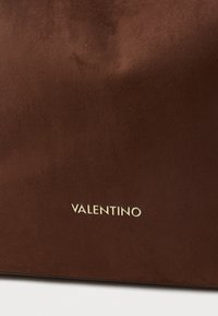 Καφέ δερμάτινη τσάντα χεριού με λεία υφή. Χρυσό λογότυπο "Valentino" στην κάτω δεξιά γωνία για λεπτομέρεια branding.