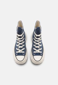 Höga sneakers i mörkblå kanvas med beige snörningar, vita gummiklädor och rundade tåskydd med svart kant.