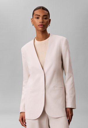 Femme aux cheveux plaqués en arrière, portant un blazer beige clair ouvert sur le devant, avec un haut et un pantalon assortis, sur fond gris.