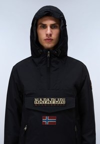 Mies pukeutunut mustaan hupulliseen takkiin, jossa on "Napapijri geographic"-etiketti ja Norjan lippu -merkki etutaskussa.