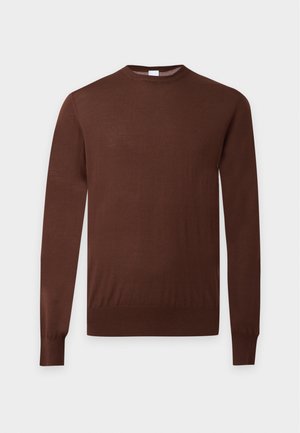 Homme portant une chemise marron à manches longues, un jean bleu clair avec une ceinture noire, et des chaussures bicolores marron et beige, debout devant un fond uni.