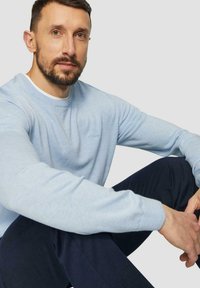 Hellesblaues Sweatshirt mit Rundhalsausschnitt aus weichem Stoff, mit gerippten Bündchen und kleinem Logo auf der Brust, kombiniert mit dunklen Hosen.