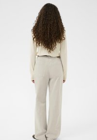 Persona ar gariem cirtainiem matiem, ģērbies krēmkrāsas džemperī un gaiši bēšās platās biksēs, stāv ar muguru pret fona.