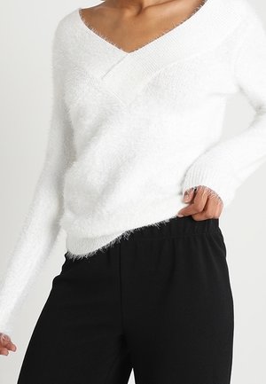 Frau trägt einen weißen, flauschigen V-Ausschnitt-Pullover und schwarze Hosen mit elastischem Bund, eine Hand hebt den Hosenbund an.