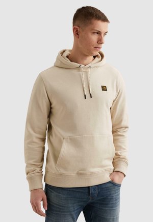 Junger Mann, der nach rechts blickt und einen beigefarbenen Kapuzenpullover mit Fronttasche und kleinem Logopatch trägt, kombiniert mit blauen Jeans, die Hände entspannt.